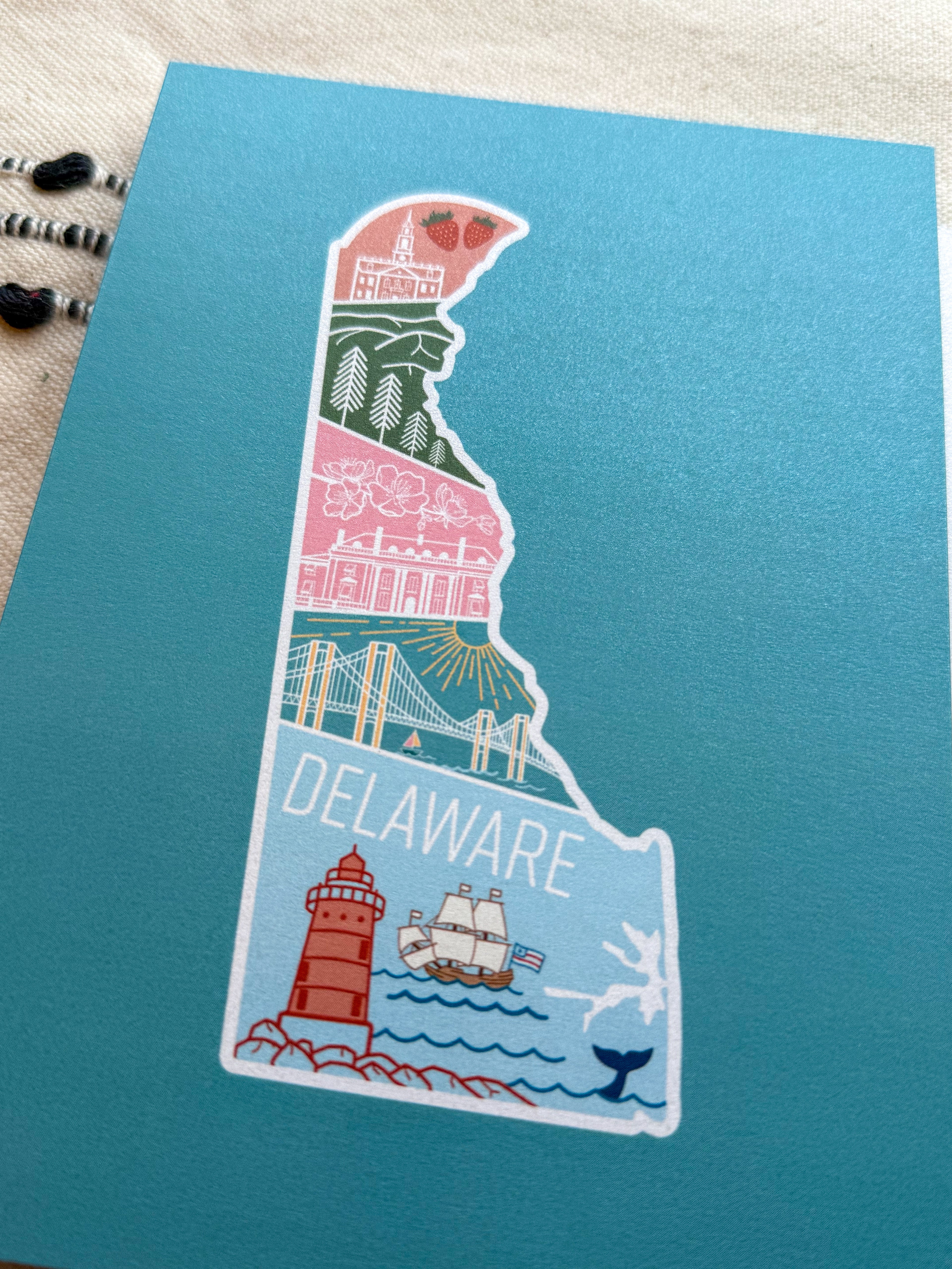 A postcard with an illustration of Delaware state in a colorful artistic style on a teal background.