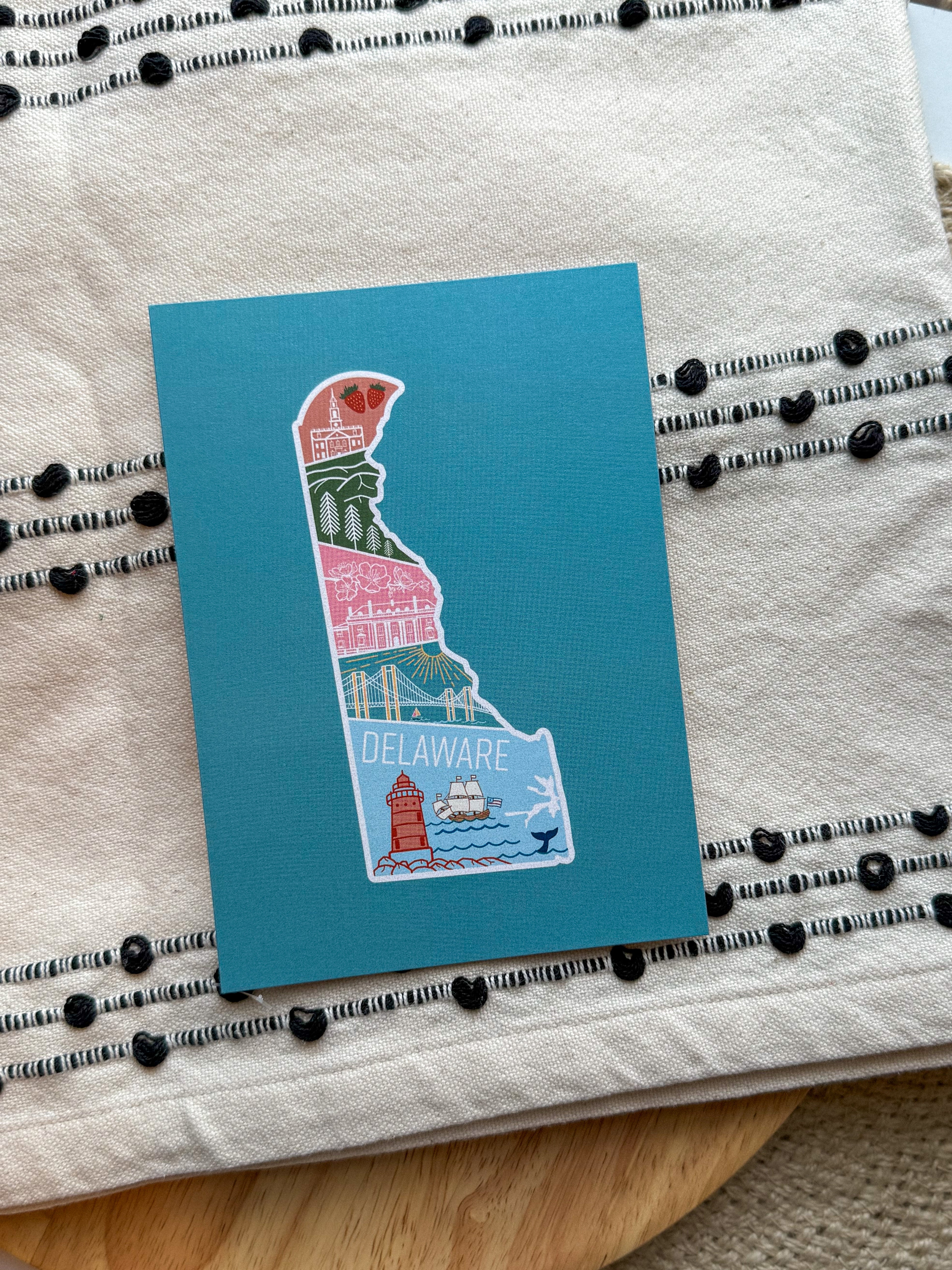 A postcard with an illustration of Delaware state in a colorful artistic style on a teal background.