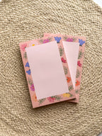 A stack of pink notepads with multicolored floral patterns on the edges, placed on a beige textured surface.