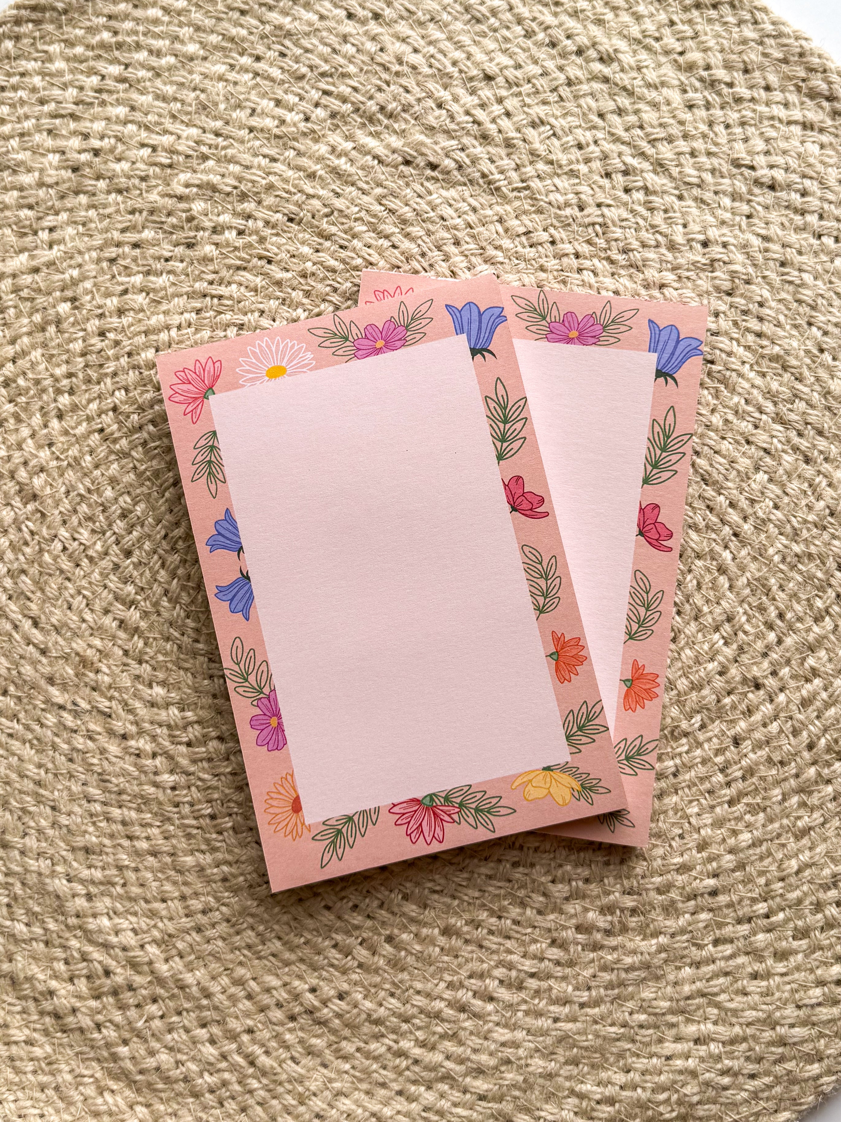 A stack of pink notepads with multicolored floral patterns on the edges, placed on a beige textured surface.