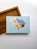 Postcard of an illustration of South Carolina state map colors and landmarks, placed on a wooden surface.