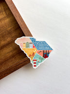 A vinyl sticker with an illustration of South Carolina state map colors and landmarks, placed on a wooden surface.