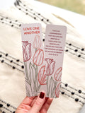 A bookmark with a floral design and a verse from the Bible John 15:12-13, held in a hand. The bookmark has a pearl paper finish with rounded corners.