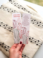 A bookmark with a floral design and a verse from the Bible John 15:12-13, held in a hand. The bookmark has a pearl paper finish with rounded corners.