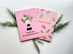 pink greeting cards with a floral illustration and text, placed on a wooden surface surrounded by various plants and flowers, with a white envelope and plastic sleeve in the background.