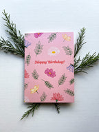 A greeting card with a floral birthday bouquet illustration and the text 'Happy Birthday!' on the front, accompanied by a white envelope, against a pink background.