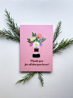 A pink thank you greeting card with a floral illustration and text, placed on a wooden surface surrounded by various plants and flowers, with a white envelope and plastic sleeve in the background.