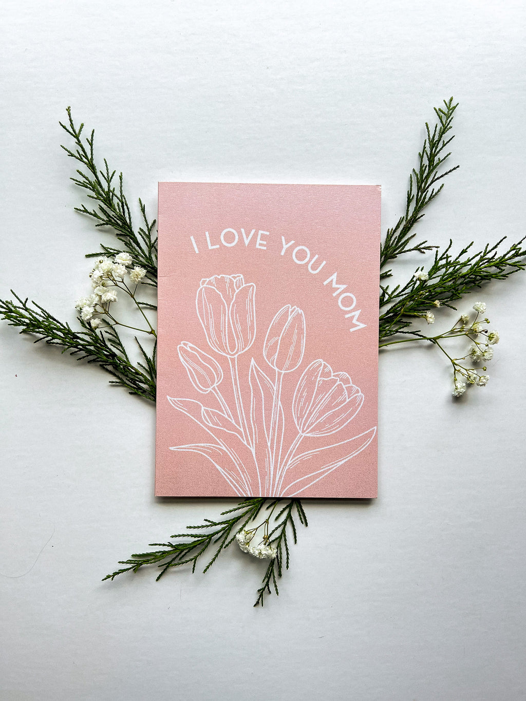 A pink greeting card with the text 'I LOVE YOU MOM' and a floral design, specifically tulips, lying on a white surface with a white envelope beside it and some foliage at the top.