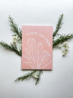 A pink greeting card with the text 'I LOVE YOU MOM' and a floral design, specifically tulips, lying on a white surface with a white envelope beside it and some foliage at the top.