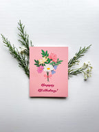 A greeting card with a floral birthday bouquet illustration and the text 'Happy Birthday!' on the front, accompanied by a white envelope, against a pink background.