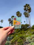Hand holding a decorative California-stamp shaped themed postcard with palm trees in the background