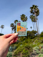 Hand holding a decorative California-stamp shaped themed postcard with palm trees in the background