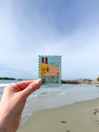 Hand holding a decorative California-stamp shaped themed postcard with beah in the background. 