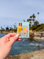 Hand holding a decorative California-stamp shaped themed postcard with palm trees in the background