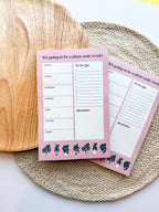 A notepad with plant illustrations at the bottom, a pink background with text areas for notes, weekly schedule. placed on a woven mat.