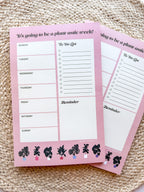 A notepad with plant illustrations at the bottom, a pink background with text areas for notes, weekly schedule. placed on a woven mat.