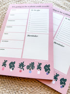 A notepad with plant illustrations at the bottom, a pink background with text areas for notes, weekly schedule. placed on a woven mat.