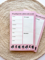 A notepad with plant illustrations at the bottom, a pink background with text areas for notes, weekly schedule. placed on a woven mat.