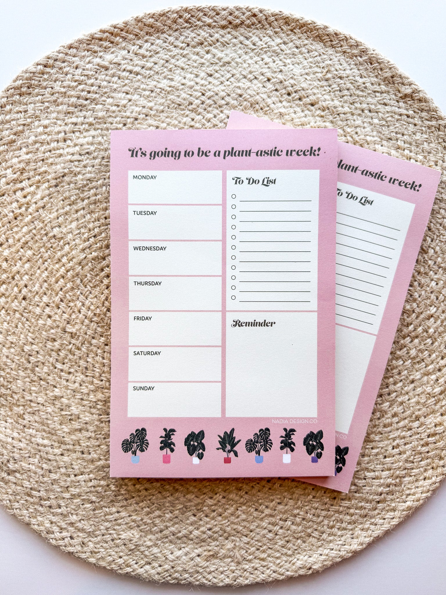 A notepad with plant illustrations at the bottom, a pink background with text areas for notes, weekly schedule. placed on a woven mat.
