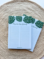 A notepad with a blue cover featuring a green monstera leaf pattern and the text 'My lists keep growing' bullet points on a beige woven surface.