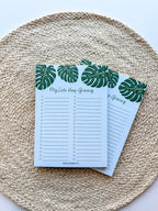 A notepad with a blue cover featuring a green monstera leaf pattern and the text 'My lists keep growing' bullet points on a beige woven surface.
