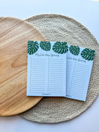 A notepad with a blue cover featuring a green monstera leaf pattern and the text 'My lists keep growing' bullet points on a beige woven surface.