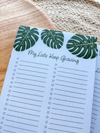 A notepad with a blue cover featuring a green monstera leaf pattern and the text 'My lists keep growing' bullet points on a beige woven surface.