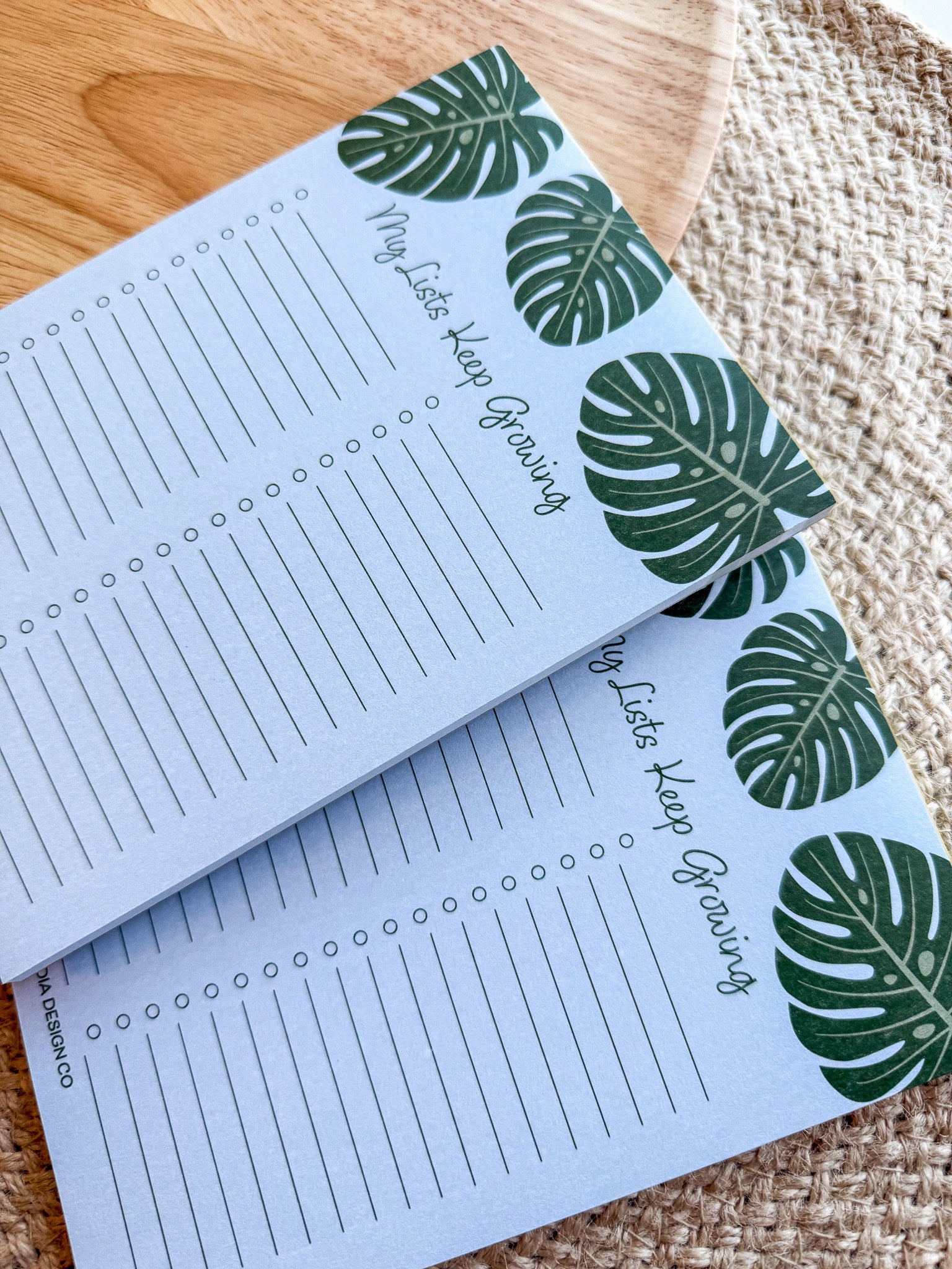 A notepad with a blue cover featuring a green monstera leaf pattern and the text 'My lists keep growing' bullet points on a beige woven surface.