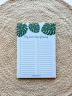 A notepad with a blue cover featuring a green monstera leaf pattern and the text 'My lists keep growing' bullet points on a beige woven surface.