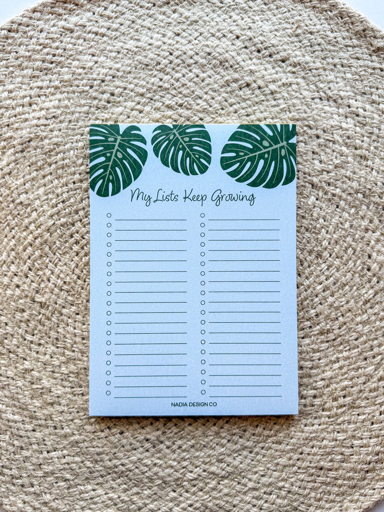 A notepad with a blue cover featuring a green monstera leaf pattern and the text 'My lists keep growing' bullet points on a beige woven surface.