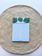 A notepad with a blue cover featuring a green monstera leaf pattern and the text 'My lists keep growing' bullet points on a beige woven surface.