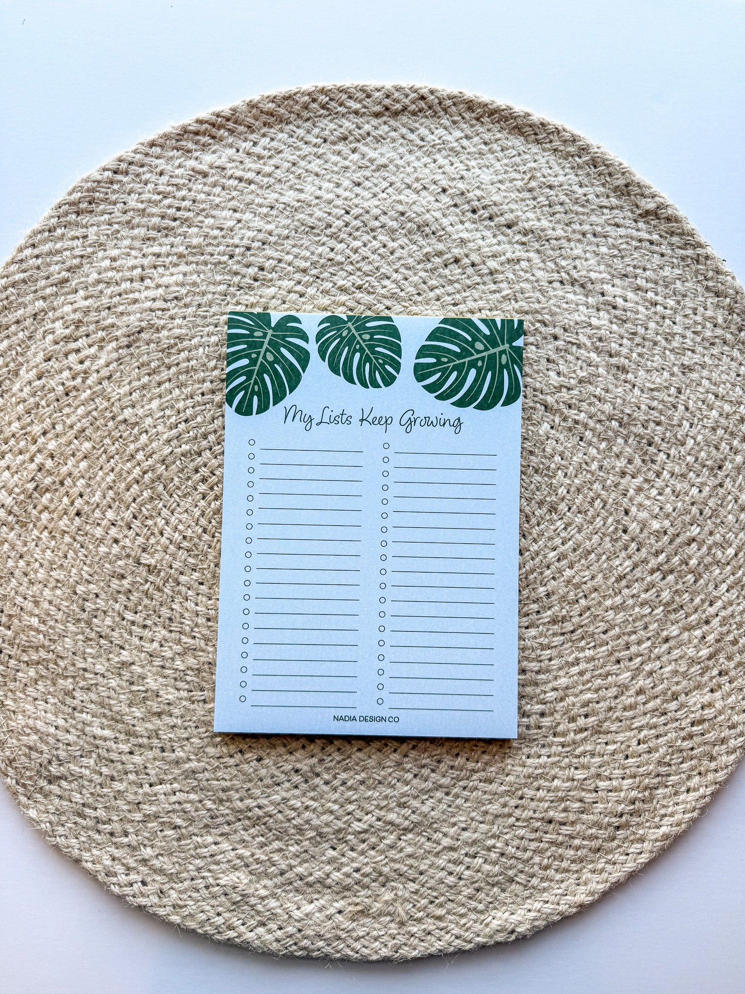 A notepad with a blue cover featuring a green monstera leaf pattern and the text 'My lists keep growing' bullet points on a beige woven surface.