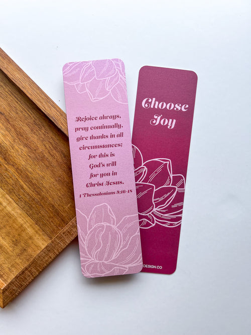 Two bookmarks with double-sided illustrations, purple, with text and floral designs, placed on a wooden surface.