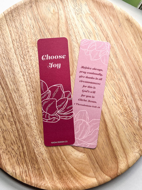 Two bookmarks with double-sided illustrations, purple, with text and floral designs, placed on a wooden surface.