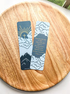 Two bookmarks with a mountain and cloud design in blue and orange colors, Psalm 25:4-5 bible verse written, placed on a wooden surface.