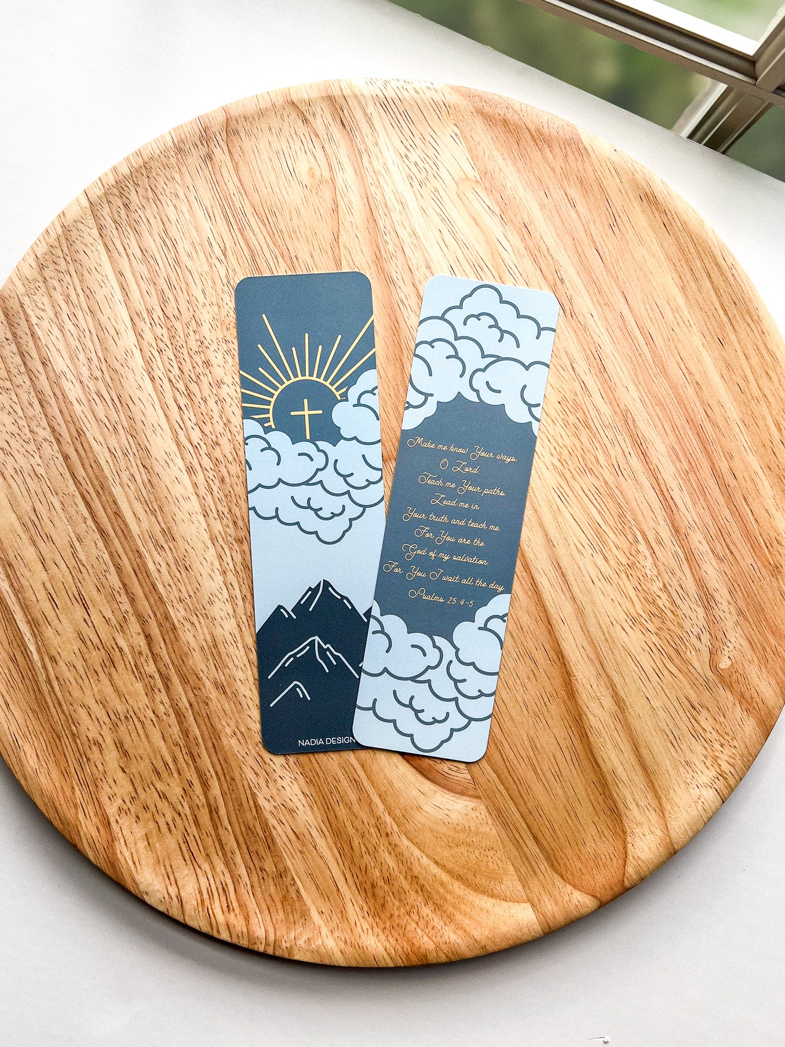 Two bookmarks with a mountain and cloud design in blue and orange colors, Psalm 25:4-5 bible verse written, placed on a wooden surface.