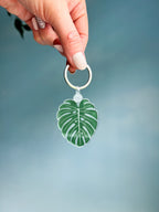 An acrylic keychain with a green leaf design, attached to a metal keyring, held by a hand against a blue background.