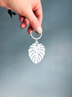 back of an acrylic keychain with a green leaf design, attached to a metal keyring, held by a hand against a blue background.
