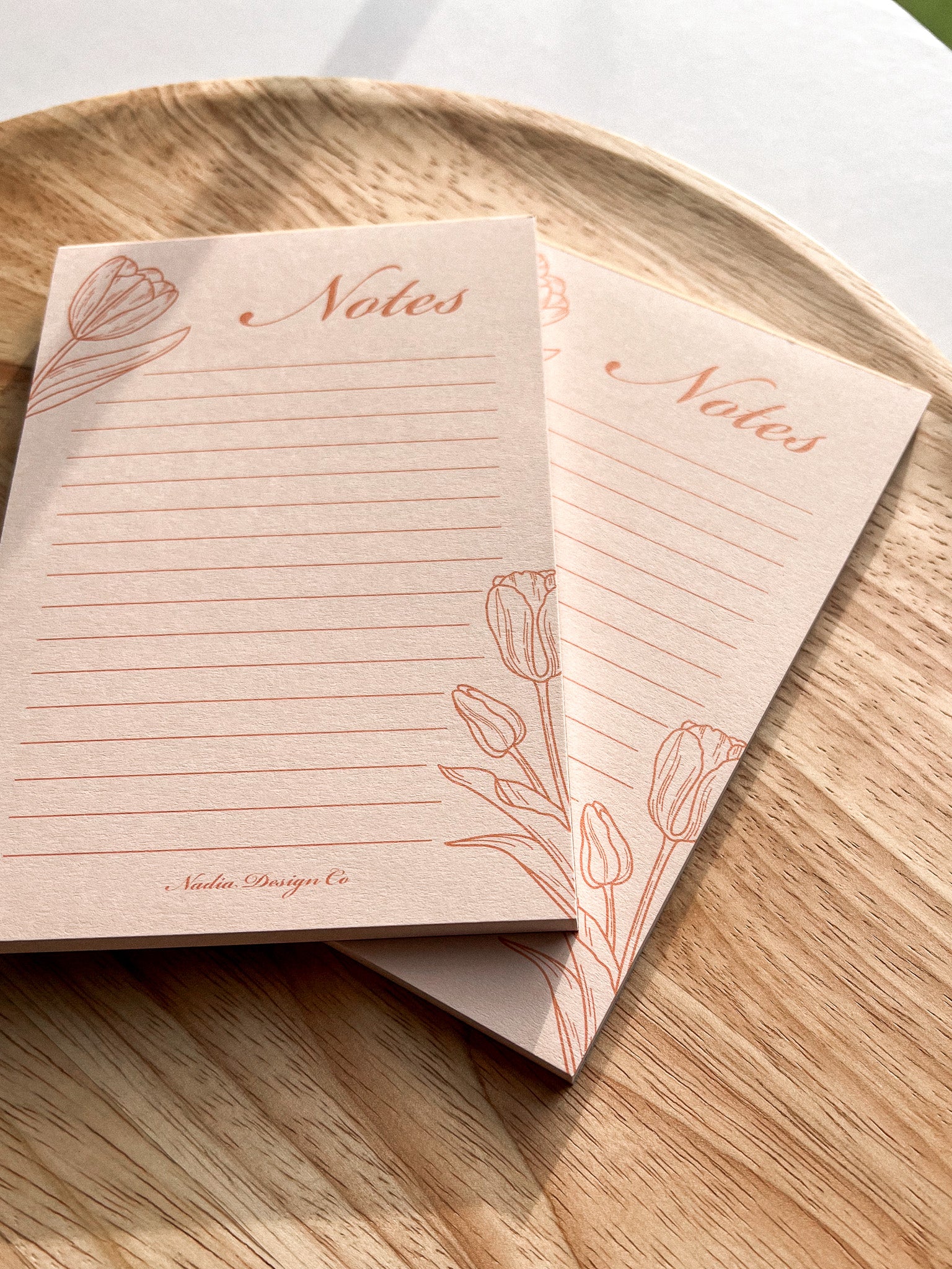 A floral tulip peachy pink lined notepad, placed on a wooden round surface