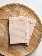 A floral tulip peachy pink notepad, placed on a wooden round surface, with wooden in the background.
