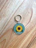 A double-sided acrylic keychain with a sunflower design on a wooden surface.