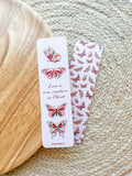 A bookmark with a white background featuring red and green butterfly illustrations and text that reads 'I am a new creation in Christ'. The bookmark is placed on a wooden surface with a patterned fabric beside it.