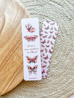 A bookmark with a white background featuring red and green butterfly illustrations and text that reads 'I am a new creation in Christ'. The bookmark is placed on a wooden surface with a patterned fabric beside it.