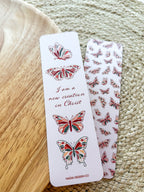 A bookmark with a white background featuring red and green butterfly illustrations and text that reads 'I am a new creation in Christ'. The bookmark is placed on a wooden surface with a patterned fabric beside it.