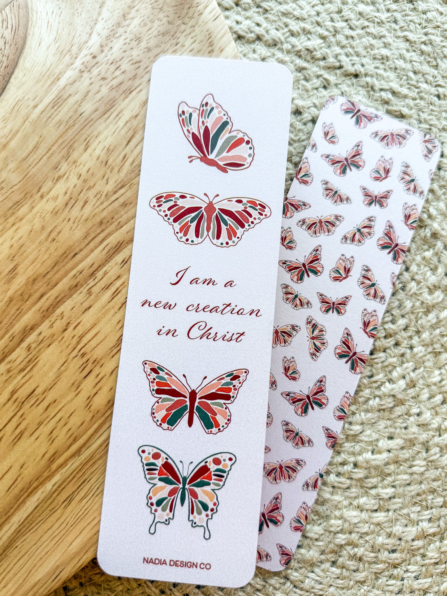 A bookmark with a white background featuring red and green butterfly illustrations and text that reads 'I am a new creation in Christ'. The bookmark is placed on a wooden surface with a patterned fabric beside it.