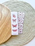 A bookmark with a double-sided illustration of butterflies, printed on pearl paper with a subtle shimmer, placed on an open book.
