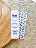 A bookmark with a butterfly illustration and a Bible verse, 2 Corinthians 5:17, laid out on a wooden surface with a patterned fabric at the top.