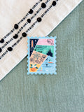 A small rectangular postage-stamp-style sticker or card with a multicolor coastal collage design, placed on a sage green textured surface next to a cream textile with black embroidered dots.