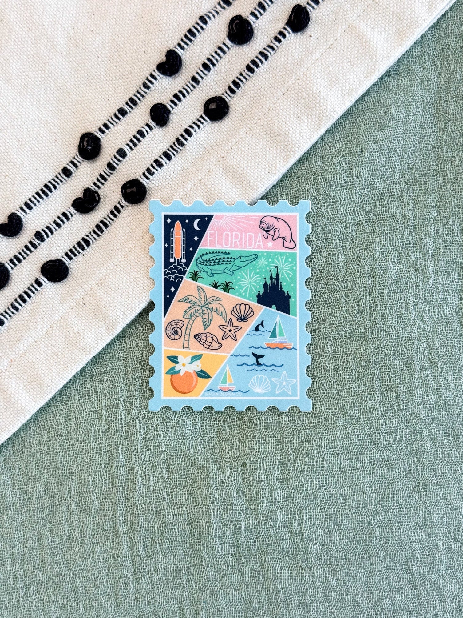 A small rectangular postage-stamp-style sticker or card with a multicolor coastal collage design, placed on a sage green textured surface next to a cream textile with black embroidered dots.