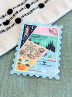 A rectangular postage-stamp-style sticker or decal with scalloped edges, featuring a pastel Florida-themed collage of palm trees, shells, a manatee, an alligator, a rocket, a castle, sailboats, and the word "FLORIDA." It is photographed on a textured light green surface with part of a cream woven textile and black beads visible at the top left.